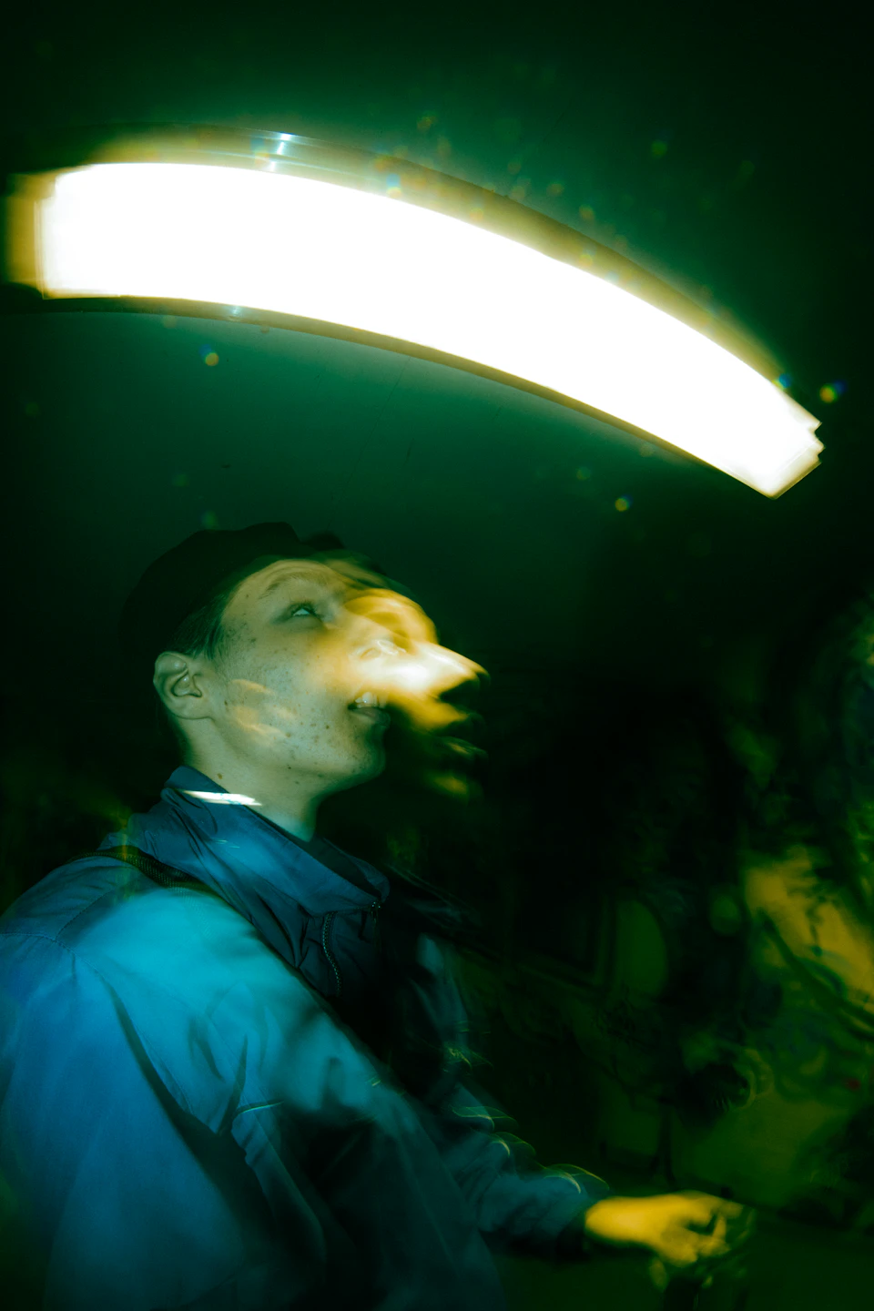 Person stands under bright light with blue jacket and black hat, looking upward