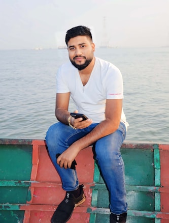 A person is seated on a boat, holding a phone, with a calm body of water in the background. The individual is wearing a white t-shirt and blue jeans, exuding a relaxed demeanor.