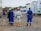 A group of construction workers, wearing blue and white protective gear with yellow and red helmets, stand on a dirt construction site. Portable buildings are visible in the background along with construction equipment.