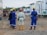 A group of construction workers, wearing blue and white protective gear with yellow and red helmets, stand on a dirt construction site. Portable buildings are visible in the background along with construction equipment.