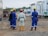 A group of construction workers, wearing blue and white protective gear with yellow and red helmets, stand on a dirt construction site. Portable buildings are visible in the background along with construction equipment.