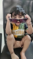A young child is sitting on a chair, holding up a pair of sunglasses with both hands. The child is wearing a multicolored shirt with the word 'Hilfiger' written across it and is gazing intently at the camera. The chair has a mesh-like design.
