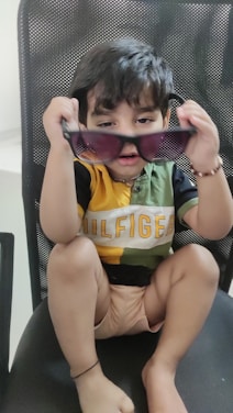 A young child is sitting on a chair, holding up a pair of sunglasses with both hands. The child is wearing a multicolored shirt with the word 'Hilfiger' written across it and is gazing intently at the camera. The chair has a mesh-like design.