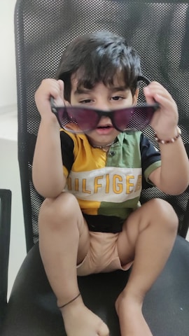 A young child is sitting on a chair, holding up a pair of sunglasses with both hands. The child is wearing a multicolored shirt with the word 'Hilfiger' written across it and is gazing intently at the camera. The chair has a mesh-like design.
