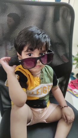 A child sits on a black mesh-backed chair, wearing oversized dark sunglasses and a colorful T-shirt with a brand logo partially visible. The child has short dark hair and is holding one side of the sunglasses with one hand. The chair and indoor setting are observable in the background.