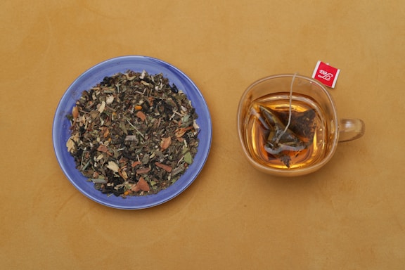 A blue plate filled with a variety of dried herbal tea ingredients is placed beside a transparent glass cup containing brewed tea with a tea bag. The background is a warm, mustard yellow.