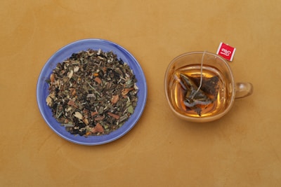 A blue plate filled with a variety of dried herbal tea ingredients is placed beside a transparent glass cup containing brewed tea with a tea bag. The background is a warm, mustard yellow.