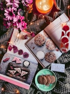 A cozy arrangement of items includes open cookbooks featuring dessert recipes, a lit candle labeled 'Mahogany,' pink flowers, a plate of cookies, and decorative lights. A handwritten note is also visible.