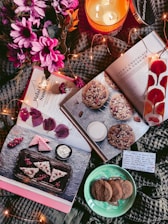 A cozy arrangement of items includes open cookbooks featuring dessert recipes, a lit candle labeled 'Mahogany,' pink flowers, a plate of cookies, and decorative lights. A handwritten note is also visible.