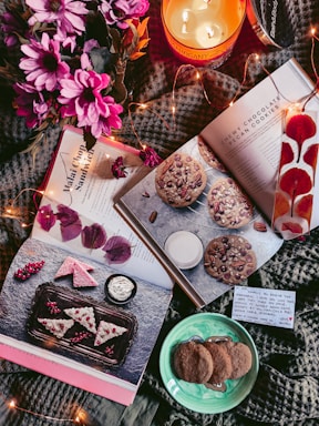 A cozy arrangement of items includes open cookbooks featuring dessert recipes, a lit candle labeled 'Mahogany,' pink flowers, a plate of cookies, and decorative lights. A handwritten note is also visible.