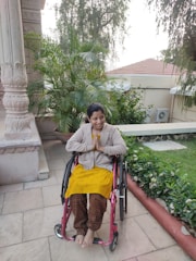 A person is seated in a wheelchair on a patio surrounded by lush green plants. The individual is smiling warmly and has their hands in a greeting or prayer gesture. The background includes architectural features and a tiled roof with trees overhead.