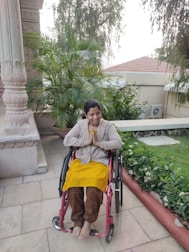 A person is seated in a wheelchair on a patio surrounded by lush green plants. The individual is smiling warmly and has their hands in a greeting or prayer gesture. The background includes architectural features and a tiled roof with trees overhead.