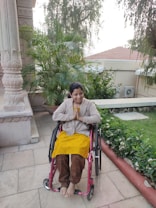 A person is seated in a wheelchair on a patio surrounded by lush green plants. The individual is smiling warmly and has their hands in a greeting or prayer gesture. The background includes architectural features and a tiled roof with trees overhead.