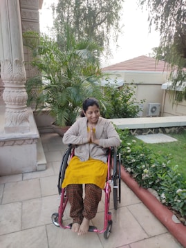 A person is seated in a wheelchair on a patio surrounded by lush green plants. The individual is smiling warmly and has their hands in a greeting or prayer gesture. The background includes architectural features and a tiled roof with trees overhead.