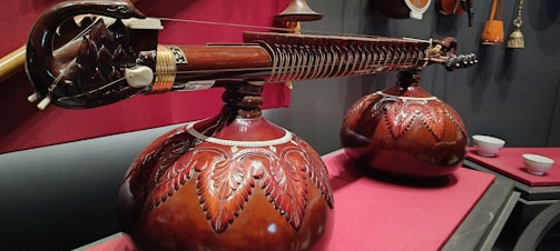 A beautifully crafted string instrument with intricate carvings rests on a red surface. The body features ornate wooden carvings with leaf-like patterns, and the strings stretch over a polished wooden surface. In the background, additional instruments and objects are visible, contributing to a rich, cultural ambiance.