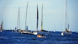 A group of sailboats and yachts floating on a blue ocean under a clear sky. Several people are visible on board, engaging in various activities. A small red and yellow kayak or rescue boat can be seen in the foreground.