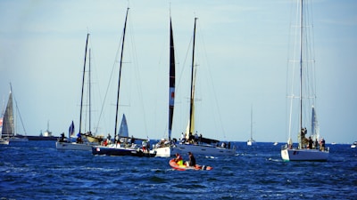 A group of sailboats and yachts floating on a blue ocean under a clear sky. Several people are visible on board, engaging in various activities. A small red and yellow kayak or rescue boat can be seen in the foreground.