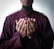 A person wearing a dark maroon robe with outstretched hands in a gesture of supplication against a neutral background.