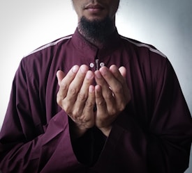 A person wearing a dark maroon robe with outstretched hands in a gesture of supplication against a neutral background.