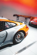 A sleek orange and silver race car with a prominent rear wing, featuring detailed alloy wheels parked indoors. Another car with red and white branding can be seen partially in the background on a pristine, tiled floor. The lighting gives a warm, vibrant ambiance with an orange gradient.