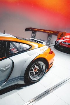 A sleek orange and silver race car with a prominent rear wing, featuring detailed alloy wheels parked indoors. Another car with red and white branding can be seen partially in the background on a pristine, tiled floor. The lighting gives a warm, vibrant ambiance with an orange gradient.
