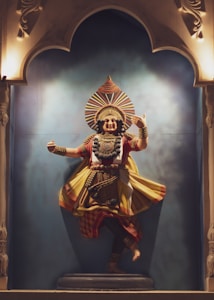A vibrant and detailed sculpture of a traditional dancer, set on a stage with ornate arch decorations. The dancer is captured in an expressive pose, wearing colorful attire with intricate patterns and textured detailing. The backdrop is a gradient blue, enhancing the focus on the figure. The sculpture exudes a sense of dynamic movement and cultural richness.