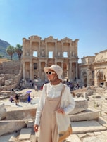 A person wearing a beige hat and sunglasses stands in front of the ancient ruins of a large stone structure with columns and arches. The backdrop features bright, clear skies and scattered tourists exploring the site. There are palm trees in the distance, and the person is holding a tote bag while dressed in light-colored clothing, appearing relaxed and contemplative.