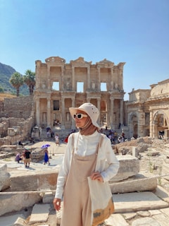 A person wearing a beige hat and sunglasses stands in front of the ancient ruins of a large stone structure with columns and arches. The backdrop features bright, clear skies and scattered tourists exploring the site. There are palm trees in the distance, and the person is holding a tote bag while dressed in light-colored clothing, appearing relaxed and contemplative.