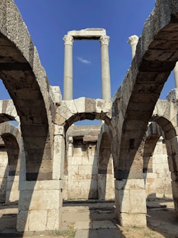 Ancient stone arches and columns stand prominently, with weathered textures and a clear blue sky in the background. The structure showcases architectural features typical of classical civilization.