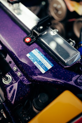 A close-up view of a car engine bay, showcasing a purple metallic component with a glossy finish and metallic flecks. There is a blue and white informational label on the surface, as well as visible bolts and brackets for securing the part. Other engine components and metallic surfaces are partially visible in the background.