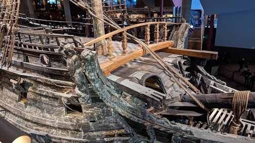 A close-up view of an old wooden ship, showcasing intricate carvings and detailed woodwork. The structure includes wooden railings, ropes, and decorative elements that highlight the craftsmanship involved. The setting appears to be indoors, possibly in a museum or exhibit space with people in the background.
