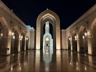 A large, elaborately decorated archway leads to a tower under a night sky. The setting is flanked by columns and arched walkways, which are illuminated by hanging lanterns. Reflections on the polished floor add to the grandeur of the scene.