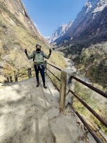 A person stands in a mountainous landscape, wearing hiking gear including a hat, mask, and backpack. The individual is on a pathway with a metal railing, overlooking a deep valley with a flowing river. Towering rocky mountains with patches of snow and green vegetation are seen in the background under a clear blue sky.