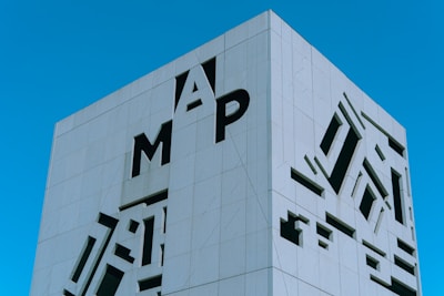 A modern building with a minimalist facade featuring large cut-out letters and abstract shapes against a clear blue sky. The building has a sleek, geometric design.