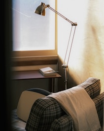 A cozy corner features a plaid armchair draped with a light blanket, positioned near a wooden desk with a rectangular tray containing papers and a pen. A sleek, metallic floor lamp stands beside the chair, casting soft light through a large frosted window, adding a warm ambiance to the room.