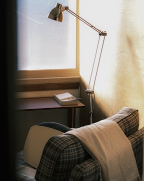 A cozy corner features a plaid armchair draped with a light blanket, positioned near a wooden desk with a rectangular tray containing papers and a pen. A sleek, metallic floor lamp stands beside the chair, casting soft light through a large frosted window, adding a warm ambiance to the room.