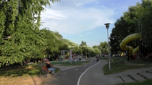 A park scene with a play area, including swings and slides, surrounded by abundant green trees. A path runs through the park with benches beside it. People are seen walking, sitting, and enjoying the playground.