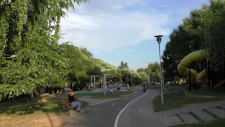 A park scene with a play area, including swings and slides, surrounded by abundant green trees. A path runs through the park with benches beside it. People are seen walking, sitting, and enjoying the playground.