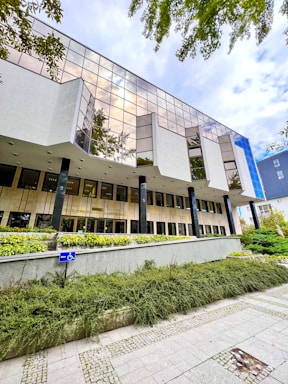 A modern building with large reflective windows and a geometric design, surrounded by greenery. The entrance is elevated and accessible by ramps, with a clearly visible handicap sign. Overhead, the sky appears cloudy, adding to an architectural feel.