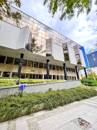 A modern building with large reflective windows and a geometric design, surrounded by greenery. The entrance is elevated and accessible by ramps, with a clearly visible handicap sign. Overhead, the sky appears cloudy, adding to an architectural feel.
