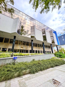 A modern building with large reflective windows and a geometric design, surrounded by greenery. The entrance is elevated and accessible by ramps, with a clearly visible handicap sign. Overhead, the sky appears cloudy, adding to an architectural feel.