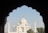 A view of the Taj Mahal framed by a dark archway silhouette, highlighting its symmetrical architecture and iconic white marble dome. The sky is clear with a few trees visible at the base of the structure.