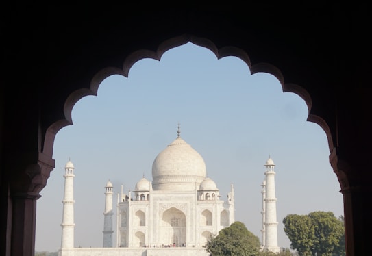 A view of the Taj Mahal framed by a dark archway silhouette, highlighting its symmetrical architecture and iconic white marble dome. The sky is clear with a few trees visible at the base of the structure.