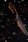 A knife with a curved blade set against a dark, snowy background. The handle appears to be crafted from a material resembling tortoiseshell, featuring a blend of brown and amber hues. The blade has a decorative pattern, with a lattice-like design near the hilt.