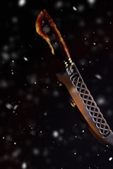 A knife with a curved blade set against a dark, snowy background. The handle appears to be crafted from a material resembling tortoiseshell, featuring a blend of brown and amber hues. The blade has a decorative pattern, with a lattice-like design near the hilt.