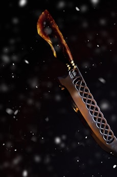 A knife with a curved blade set against a dark, snowy background. The handle appears to be crafted from a material resembling tortoiseshell, featuring a blend of brown and amber hues. The blade has a decorative pattern, with a lattice-like design near the hilt.
