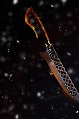 A knife with a curved blade set against a dark, snowy background. The handle appears to be crafted from a material resembling tortoiseshell, featuring a blend of brown and amber hues. The blade has a decorative pattern, with a lattice-like design near the hilt.