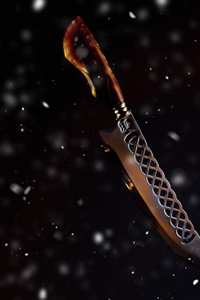 A knife with a curved blade set against a dark, snowy background. The handle appears to be crafted from a material resembling tortoiseshell, featuring a blend of brown and amber hues. The blade has a decorative pattern, with a lattice-like design near the hilt.