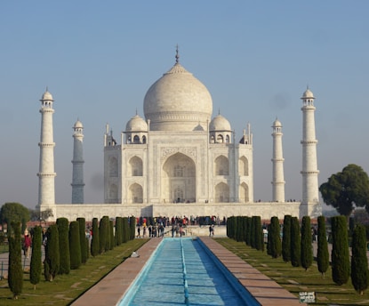 A large white marble mausoleum with a central dome and four symmetrical minarets stands majestically at the center. In front, a long reflective pool runs through a well-maintained garden with evenly spaced trees. A clear blue sky forms the backdrop as a crowd of people gathers near the entrance.