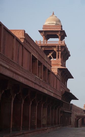 A historic architectural structure featuring intricate carvings and columns, with a prominent domed tower rising above. The building is made of red stone and exhibits traditional design elements, showcasing a blend of arches and balconies.